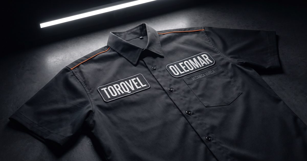 Camisa de uniforme de oficina, cinza-escura, estendida sobre concreto polido, com dois bordados grandes de marcas de fornecedor inventadas, TORQVEL e OLEOMAR, ocupando o peito, e o nome da própria oficina, MECANICA VARELA, bordado bem pequeno acima do bolso